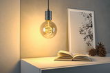 Hängende Glühbirne mit warmweißem LED-Filament über weißem Tisch mit Buch und Kiefernzapfen.