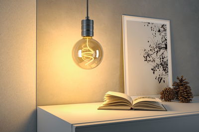 Hängende Glühbirne mit warmweißem LED-Filament über weißem Tisch mit Buch und Kiefernzapfen.