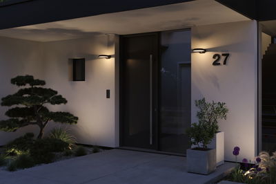 Modern outdoor lighting with black wall lamp on white facade by front door and plants at night.