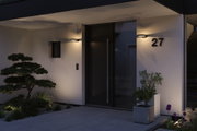 Moderne Außenbeleuchtung mit schwarzer Wandlampe an weißer Hausfassade neben Haustür und Pflanzen.