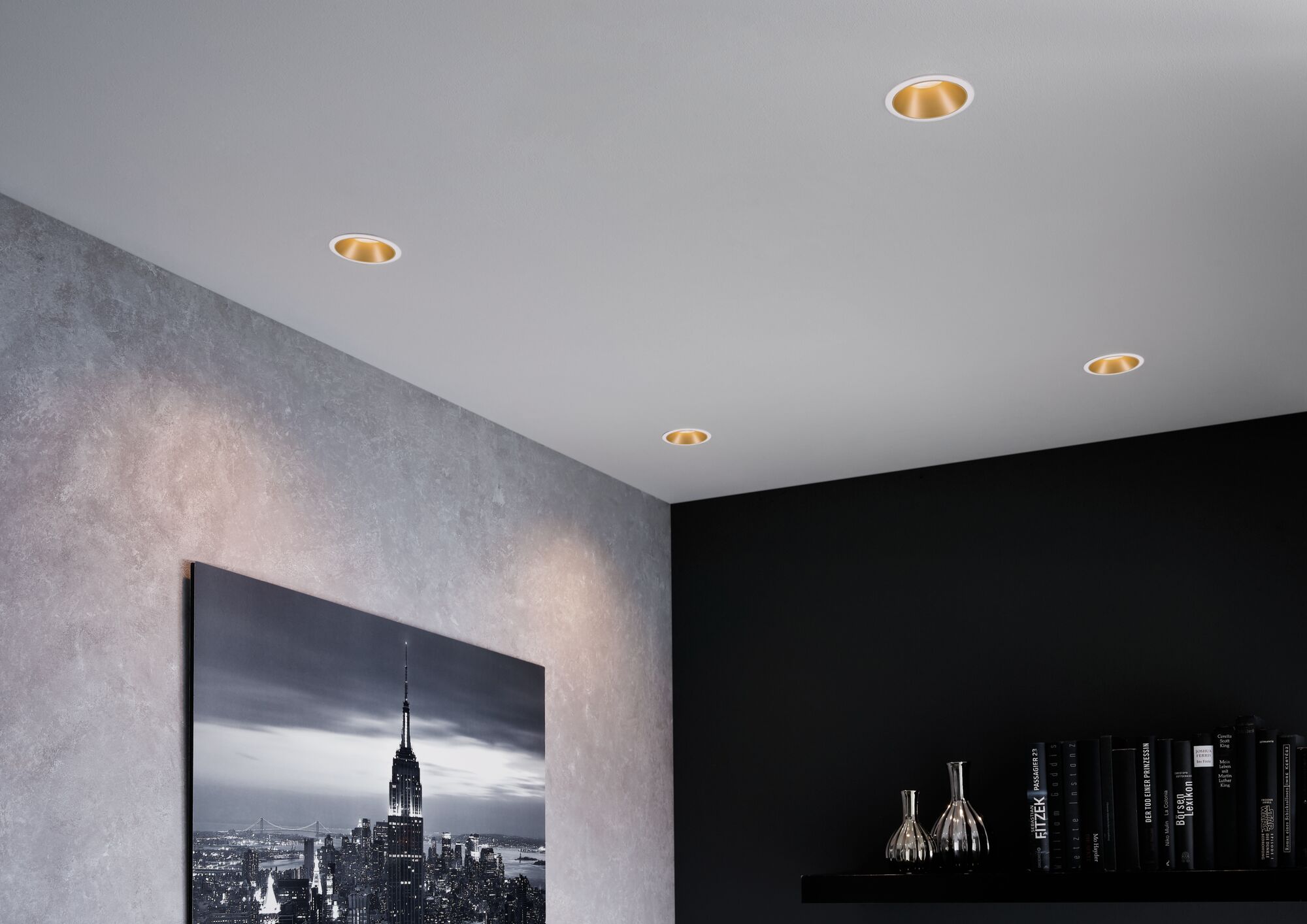 Four white recessed spotlights with gold reflector in modern living room with cityscape photo and bookshelf.