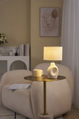 Beige table lamp with fabric shade and ceramic base on gold side table in cozy living room.