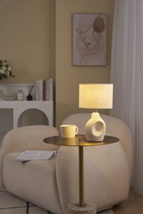 Beige Tischlampe mit Stoffschirm und Keramikfuß auf goldfarbenem Beistelltisch im Wohnzimmer.