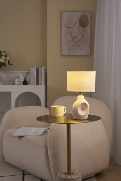Beige Tischlampe mit Stoffschirm und Keramikfuß auf goldfarbenem Beistelltisch im Wohnzimmer.