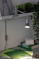 Grijze LED-klemspot met flexibele arm aan plank, verlicht groen knutselpapier op bureau met schaar.