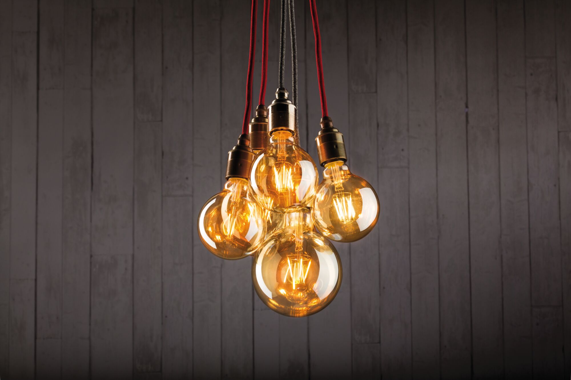 Five hanging warm light bulbs with red and black cords against a grey wooden wall background.