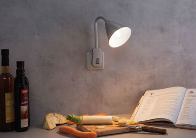 Grijze wandlamp met flexibele arm verlicht keukenblad met groenten en open kookboek.