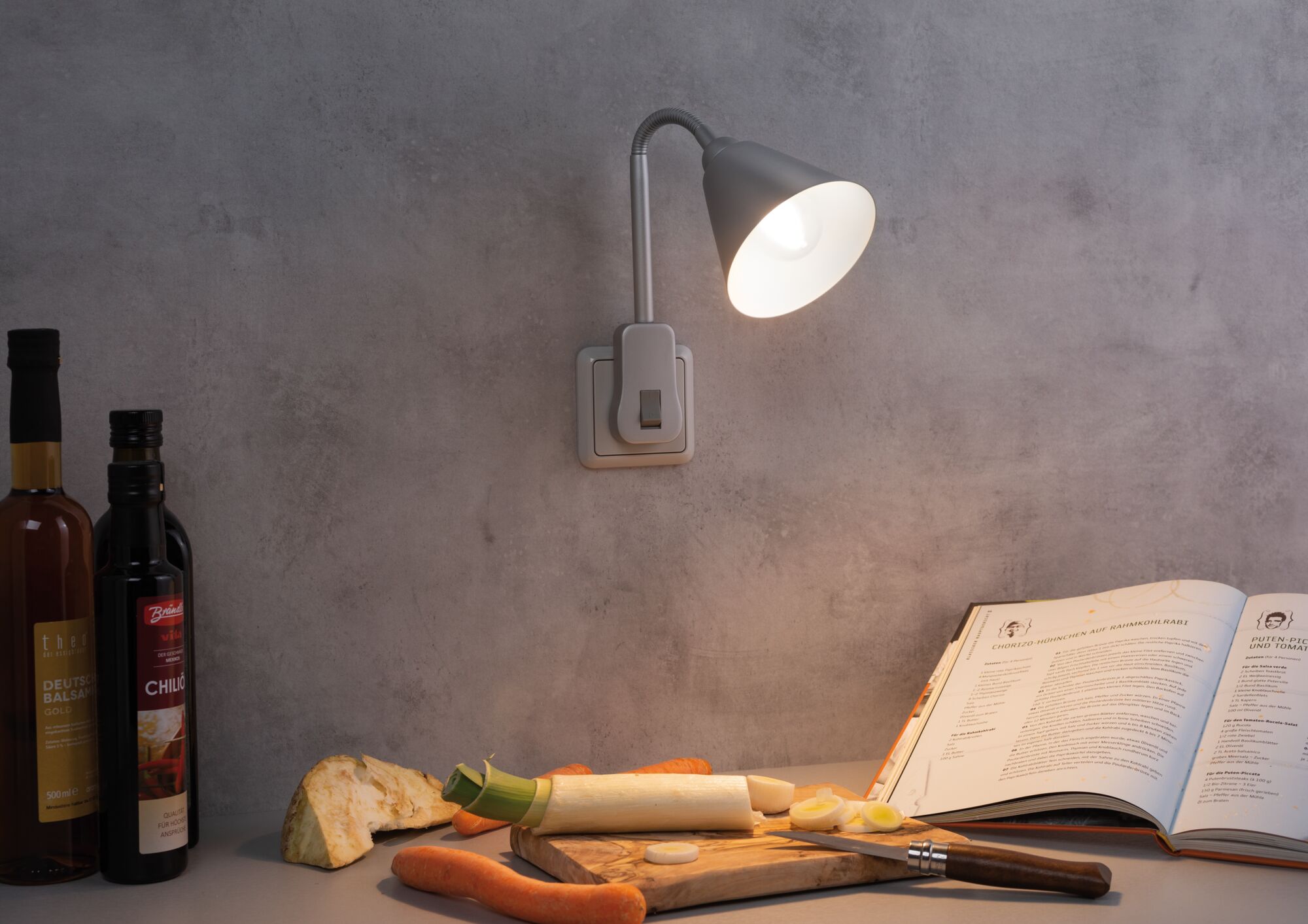 Grijze wandlamp met flexibele arm verlicht keukenblad met groenten en open kookboek.