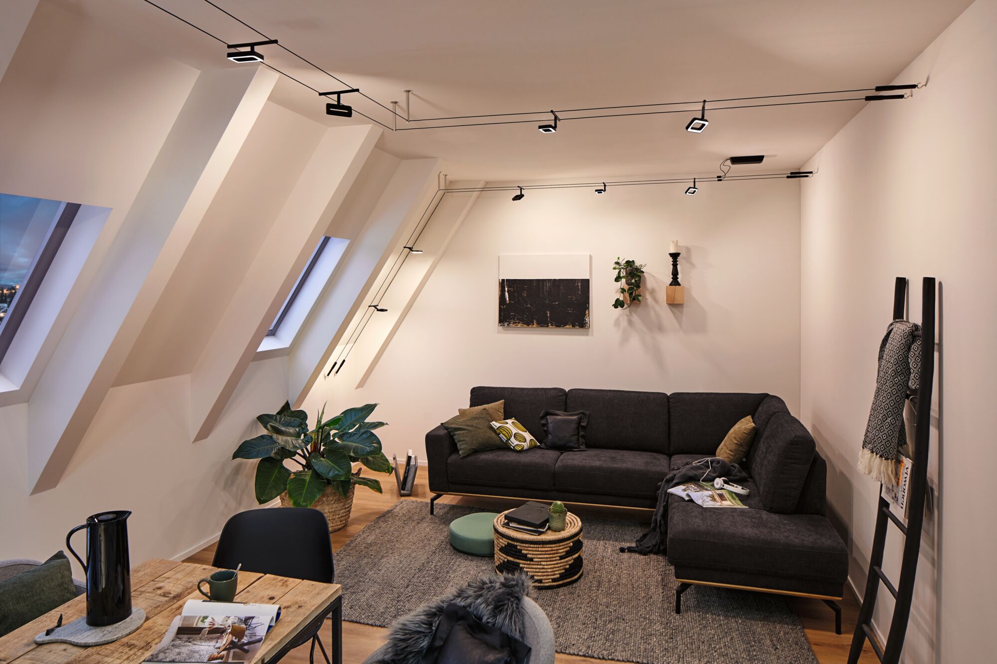 Modernes Wohnzimmer mit schwarzem Stoffsofa, grauem Teppich und Paulmann LED-Seilzugbeleuchtung an der Decke.