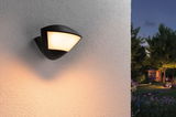 Black outdoor wall light with warm white glow on white wall, garden visible in dusk background