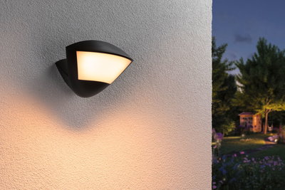 Black outdoor wall light with warm white glow on white wall, garden visible in dusk background