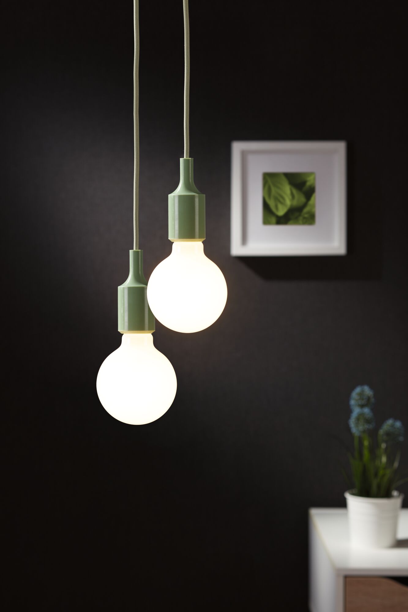 Zwei hängende LED-Glühbirnen mit grünen Fassungen vor schwarzer Wand und weißem Tisch mit Pflanze.