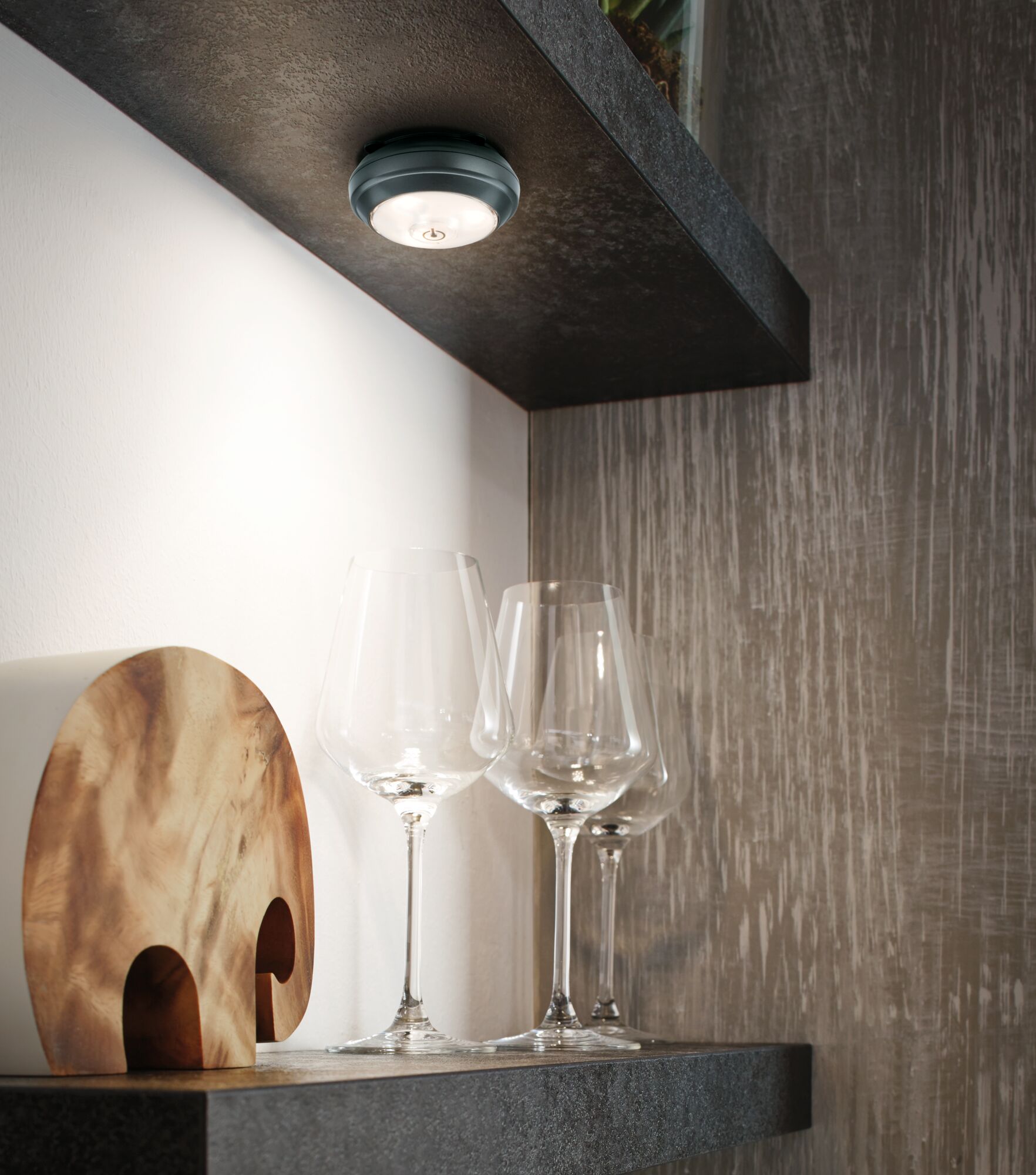 Round black LED ceiling light with warm white glow above clear wine glasses on dark shelf.