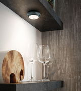 Round black LED ceiling light with warm white glow above clear wine glasses on dark shelf.