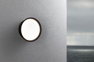 Round outdoor wall light with black frame and white diffuser on grey wall overlooking sea