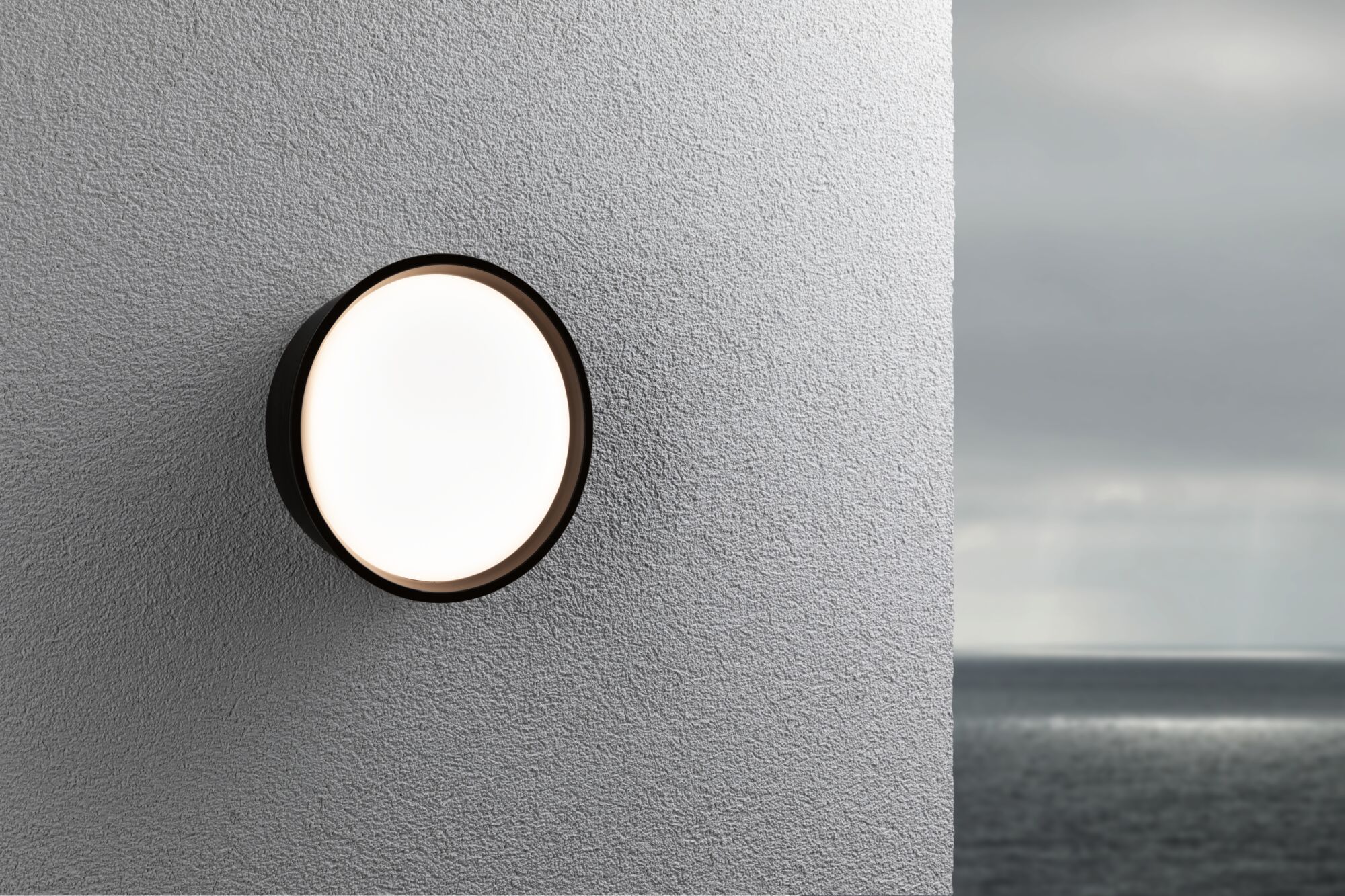 Runde Außenwandleuchte mit schwarzem Rahmen und weißem Diffusor an grauer Wand mit Meerblick