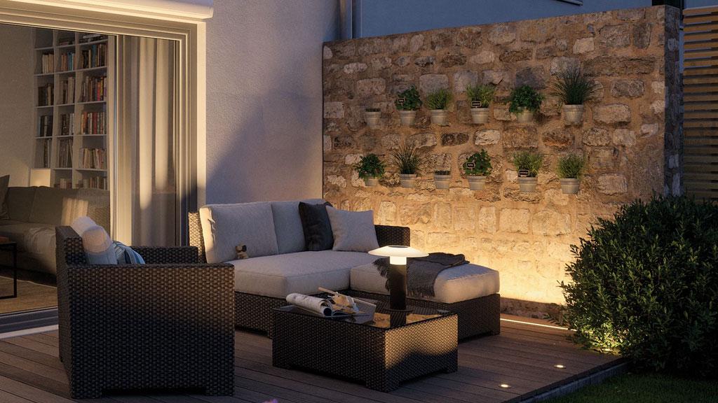 Hyggelig terrasse med grå rattan lounge og varm hvid LED-havebelysning på naturstensmur.