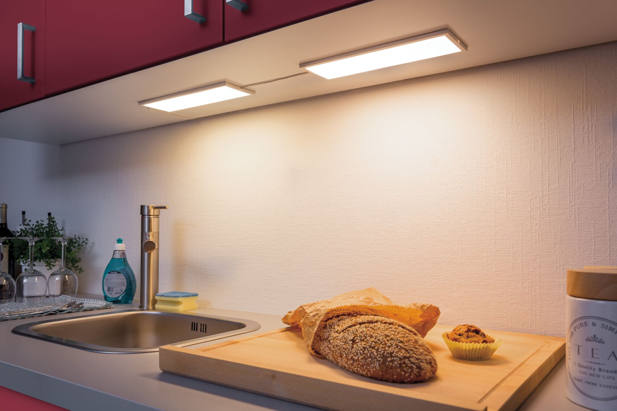 Plan de travail de cuisine avec évier en acier inoxydable, planche à découper en bois, pain et éclairage LED sous meuble blanc chaud.