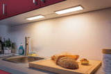 Plan de travail de cuisine avec évier en acier inoxydable, planche à découper en bois, pain et éclairage LED sous meuble blanc chaud.