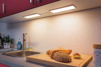 Plan de travail de cuisine avec évier en acier inoxydable, planche à découper en bois, pain et éclairage LED sous meuble blanc chaud.