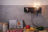 Schwarze Wandlampe mit Glühbirne und Ablagefläche über Küchenarbeitsplatte mit Paprika und Kräutern