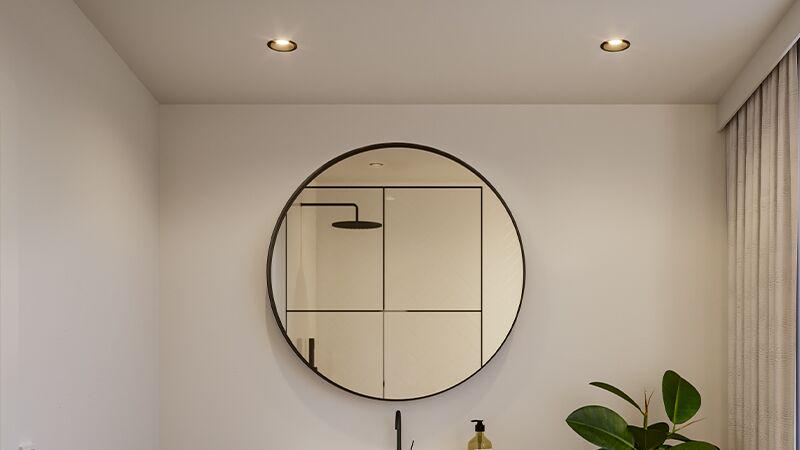 Round bathroom mirror with black frame and two warm white recessed lights in damp room ceiling.