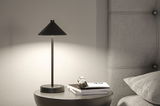 Lampe de table noire en métal avec abat-jour conique sur table de chevet grise près d’une tête de lit rembourrée.