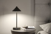 Schwarze Tischlampe aus Metall mit konischem Schirm auf grauem Nachttisch neben gepolstertem Kopfteil.