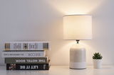Crème witte keramische tafellamp met stoffen kap naast boeken en kleine groene plantenpot.