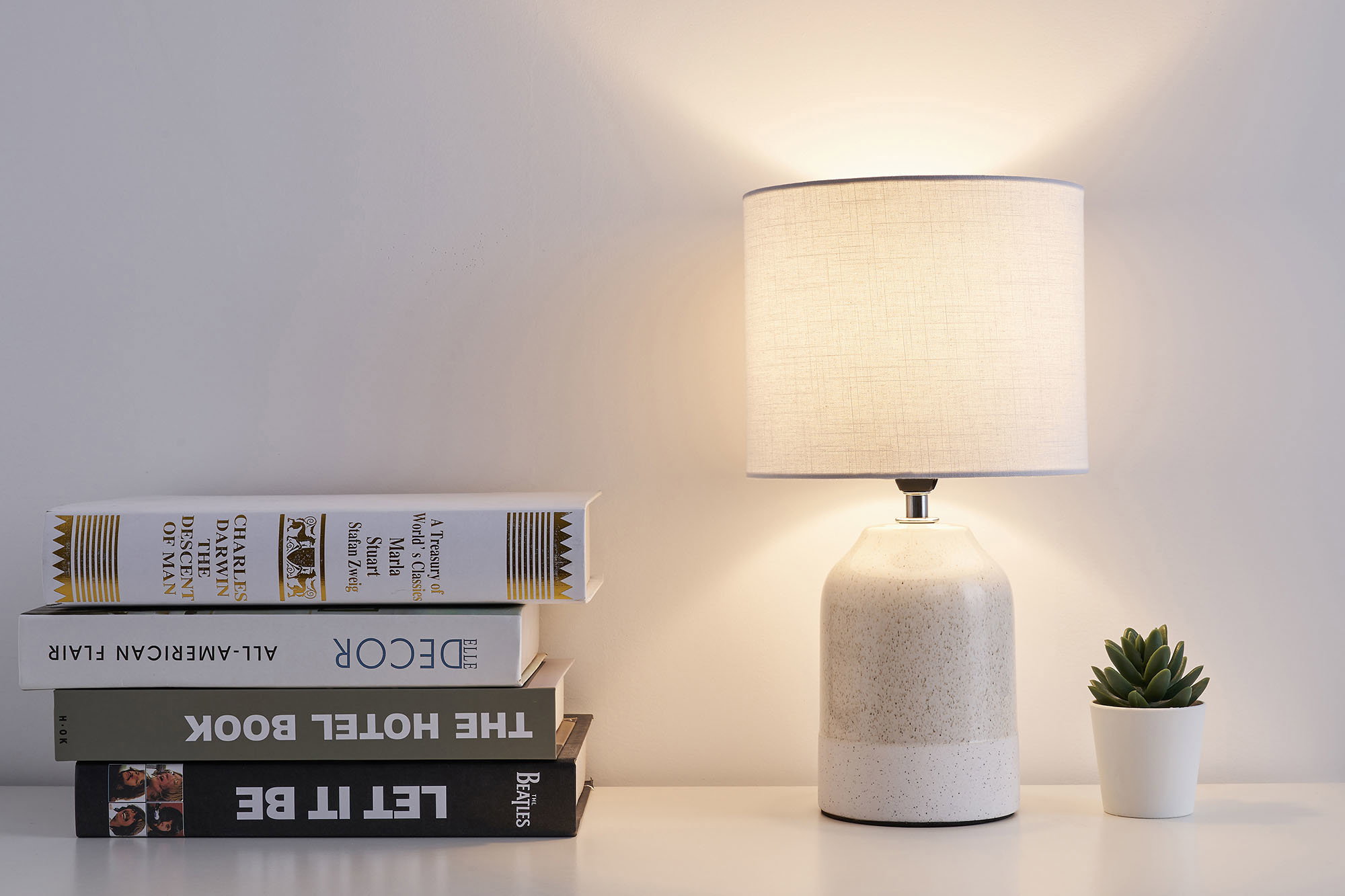 Cream ceramic table lamp with light fabric shade beside books and small green succulent pot