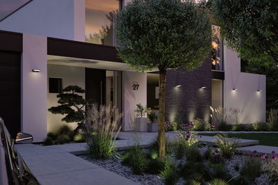 Modern house facade with white aluminum wall lights illuminating garden with plants and tree.
