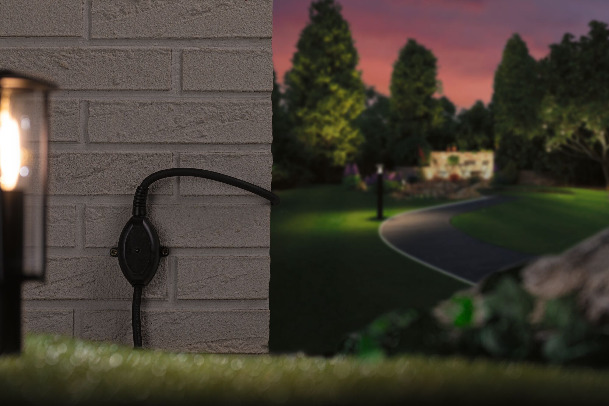 Zwarte buitenstekker op grijze bakstenen muur met verlicht tuinpad op de achtergrond bij schemering