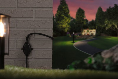 Zwarte buitenstekker op grijze bakstenen muur met verlicht tuinpad op de achtergrond bij schemering