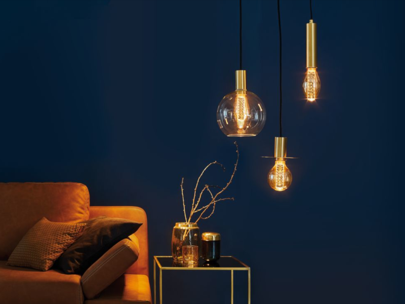 Drei goldene Pendelleuchten mit warmem Licht vor dunkelblauer Wand neben braunem Sofa und Beistelltisch.