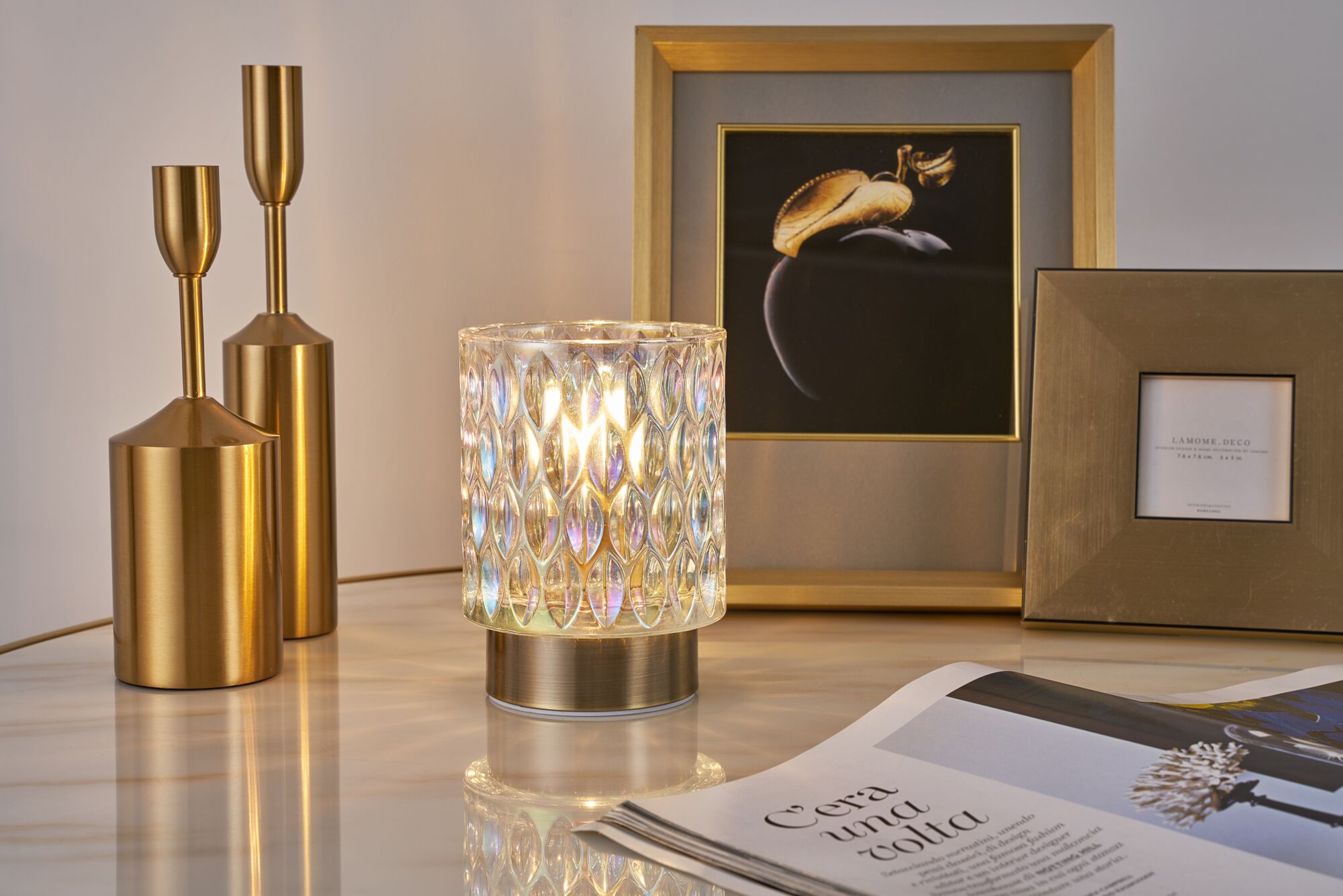 Decoratieve tafellamp met gestructureerd glas en gouden metalen voet naast gouden kandelaren en fotolijsten