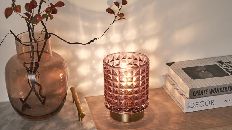 Lampe de table mobile en verre rose avec socle doré sur surface en bois près d'un vase et des livres.