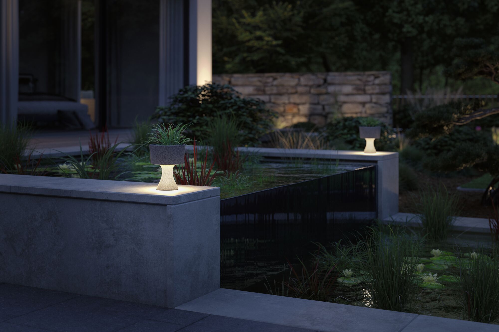 Verlichte grijze stenen plantenbak op betonnen muur in tuin met vijver en waterplanten bij schemering.