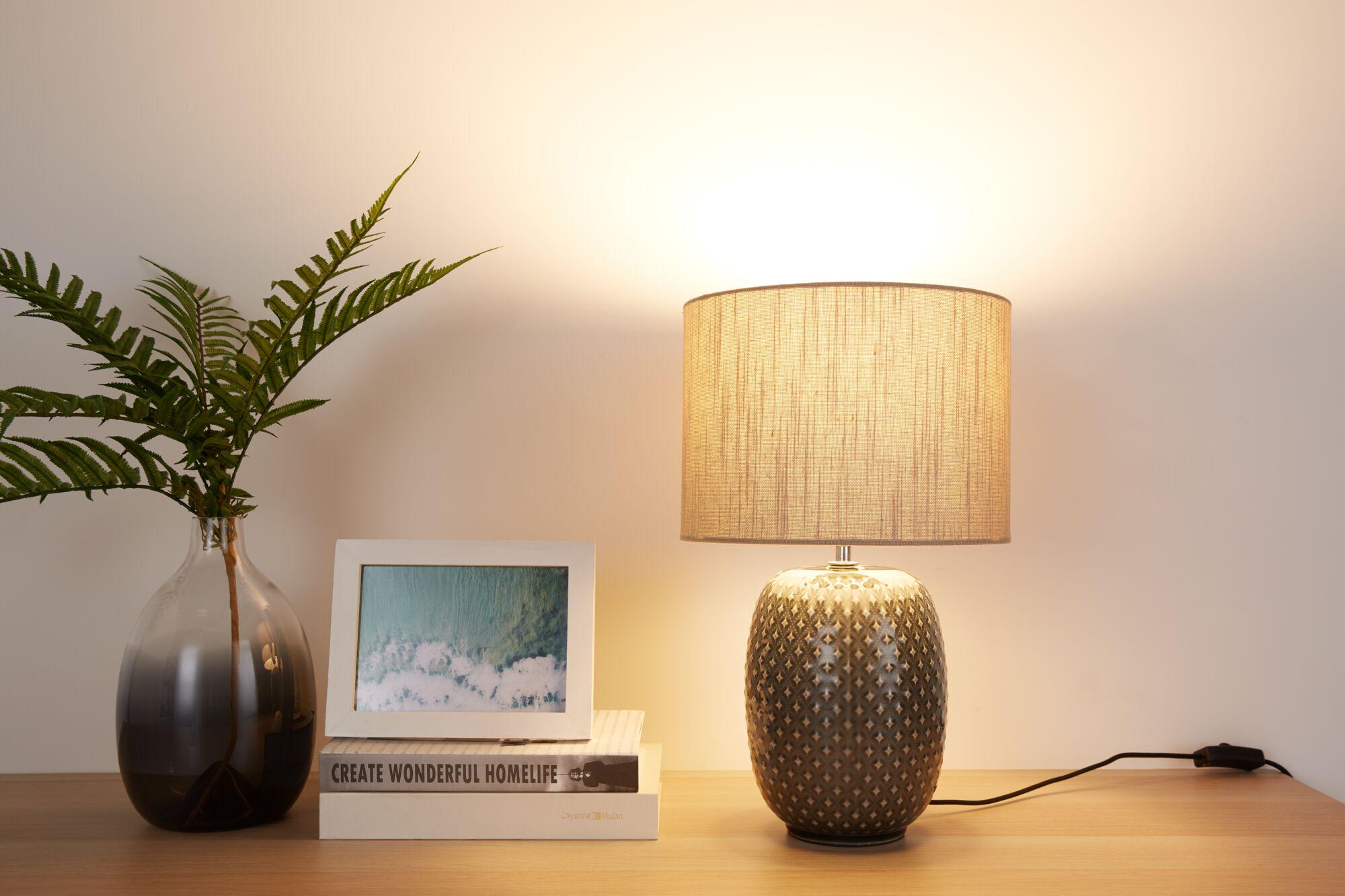 Keramisk bordlampe med beige stofskærm på træbord ved siden af vase med grønne blade og billedramme