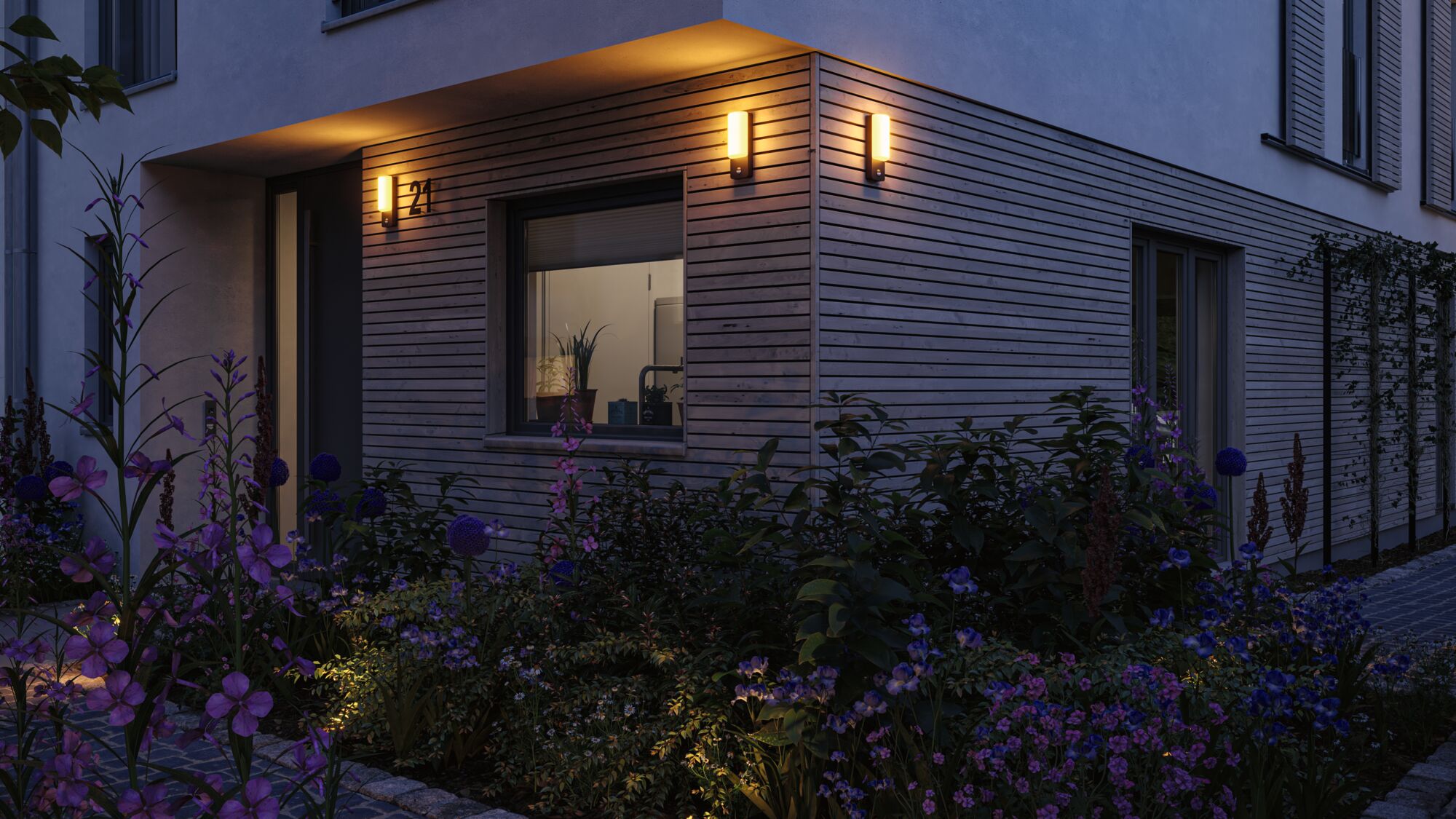 Modern outdoor wall lights with warm white light on wooden facade illuminating garden with purple flowers at dusk