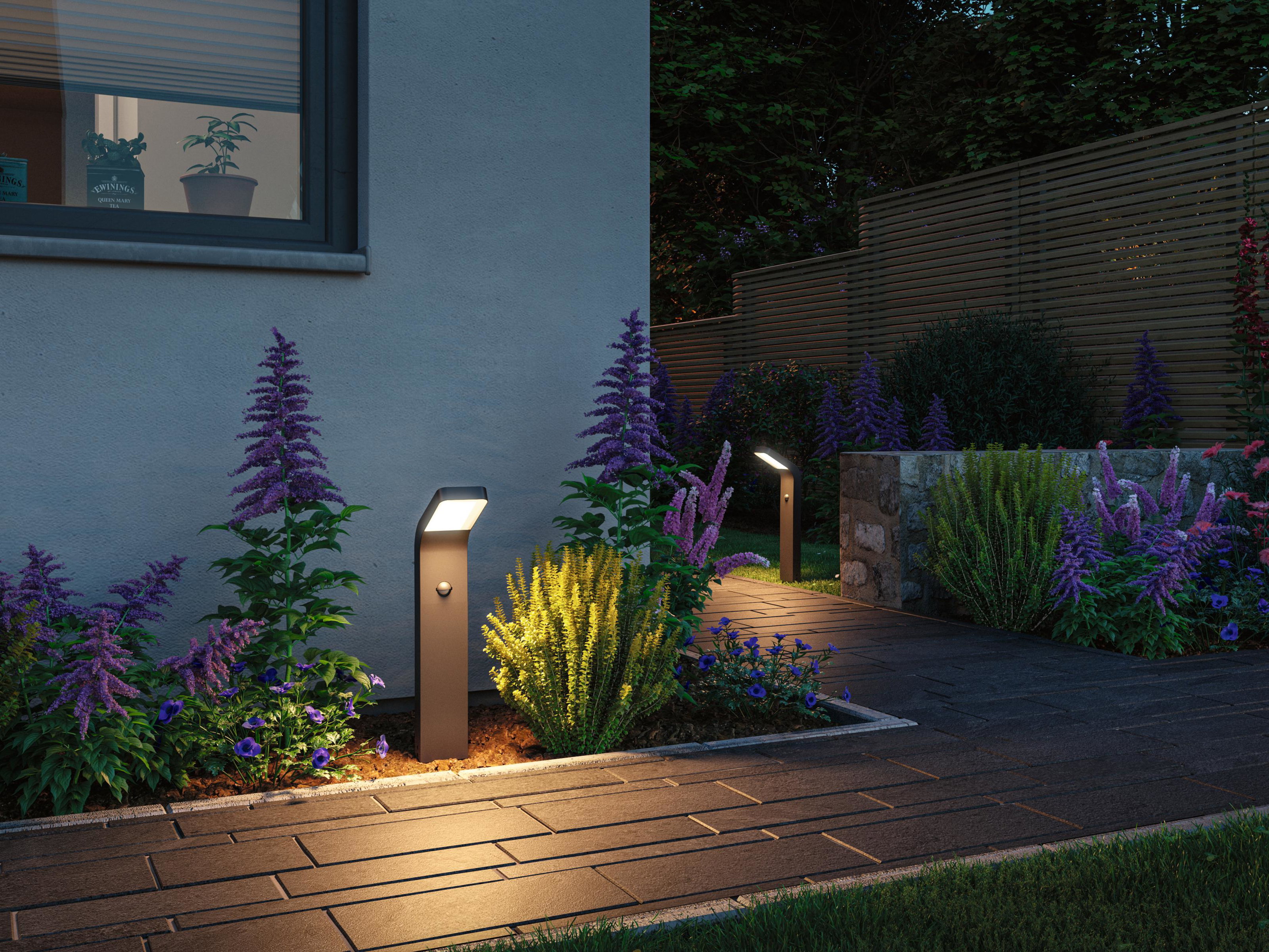 Moderne braune LED-Wegeleuchte aus Metall beleuchtet Gartenweg mit bunten Blumen bei Dämmerung