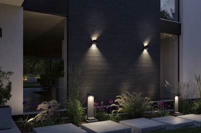 Modern outdoor lighting with grey bollard lights and wall lamps on black brick wall in garden.