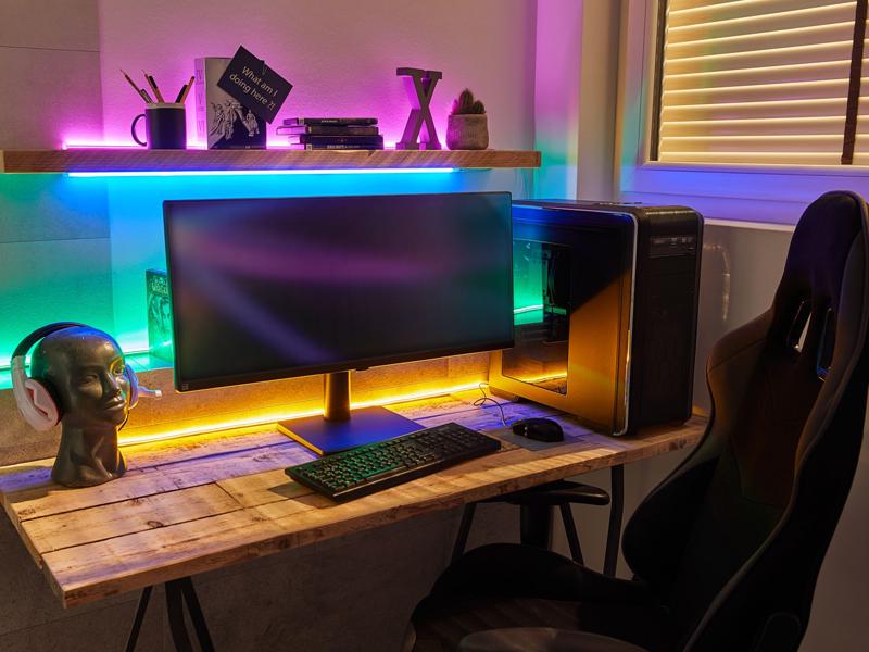 Colorful LED USB strip lighting behind monitor and shelf on wooden desk with PC and gaming chair for ambient light.
