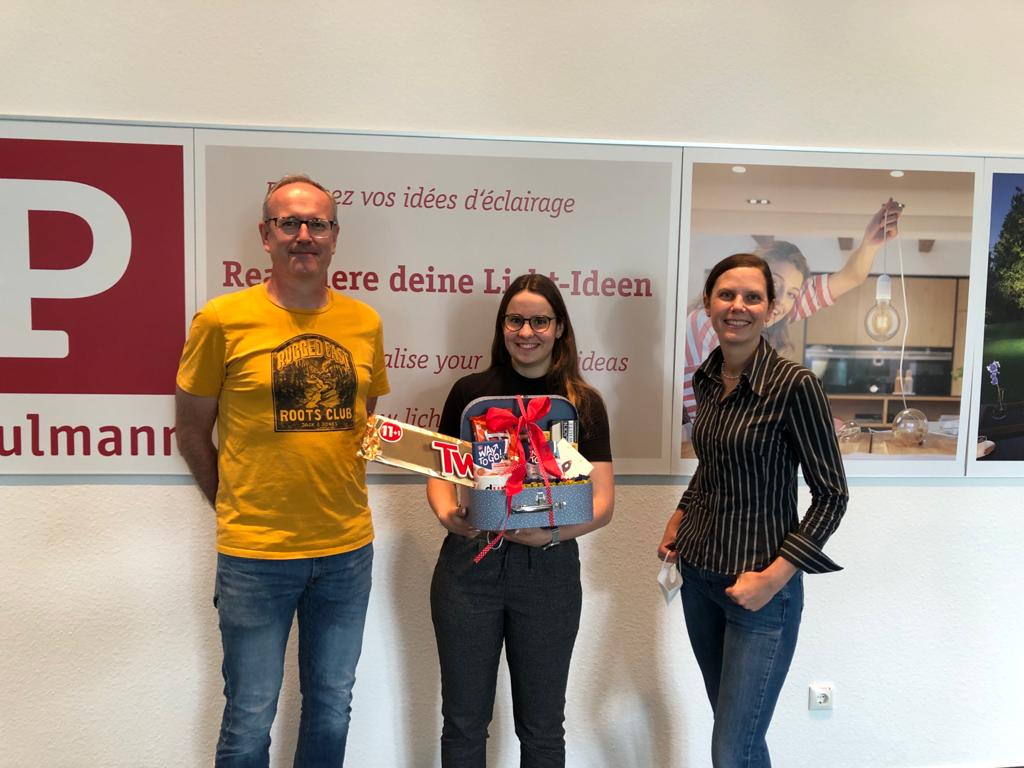 Three people standing in front of Paulmann sign, one holding gift box with sweets, friendly setting