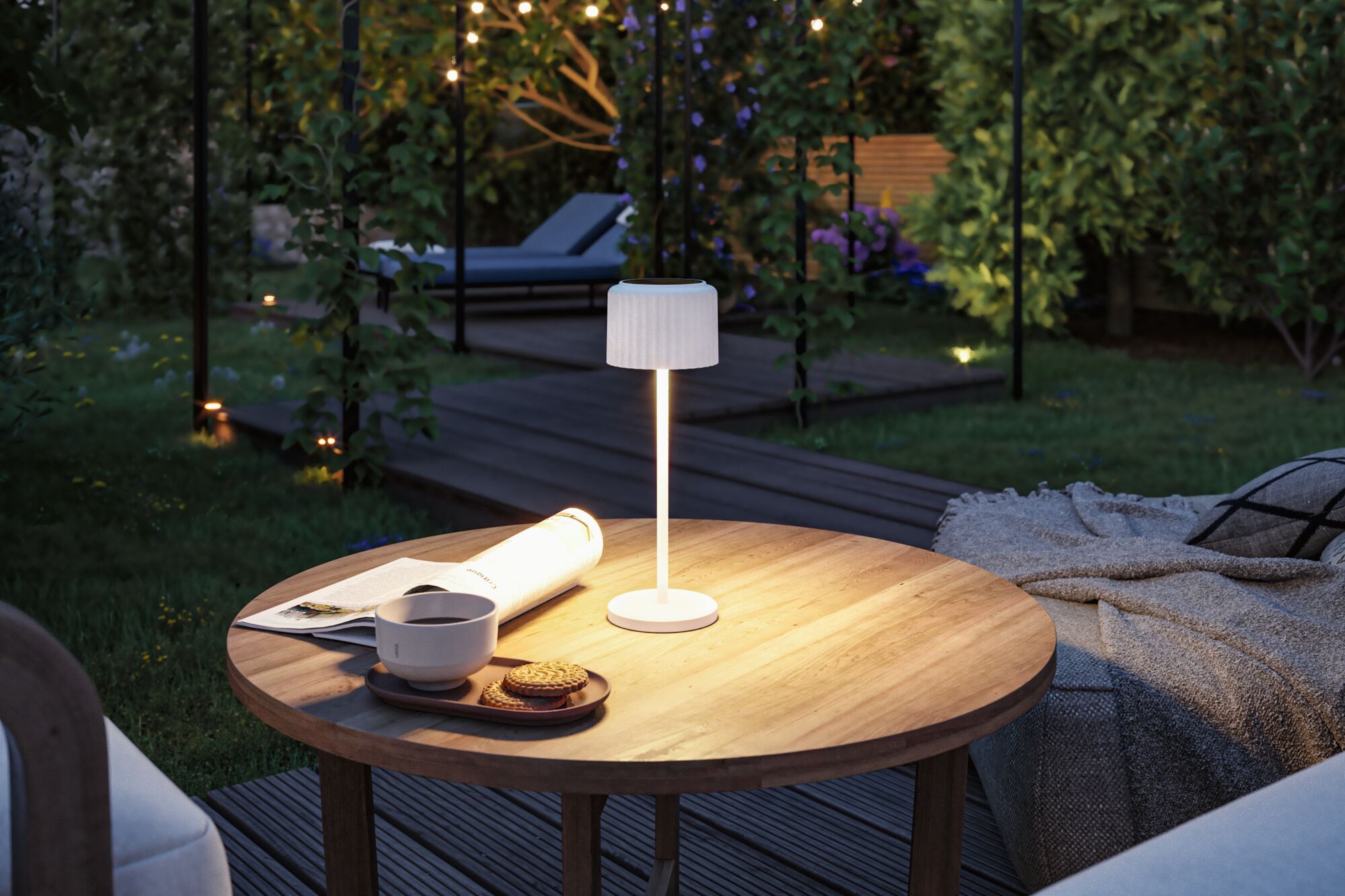 White metal LED table lamp on wooden garden table with cup, cookies, and newspaper in evening light