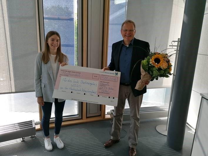 Zwei Personen in Büro halten großen Scheck und Blumenstrauß bei Spendenübergabe.