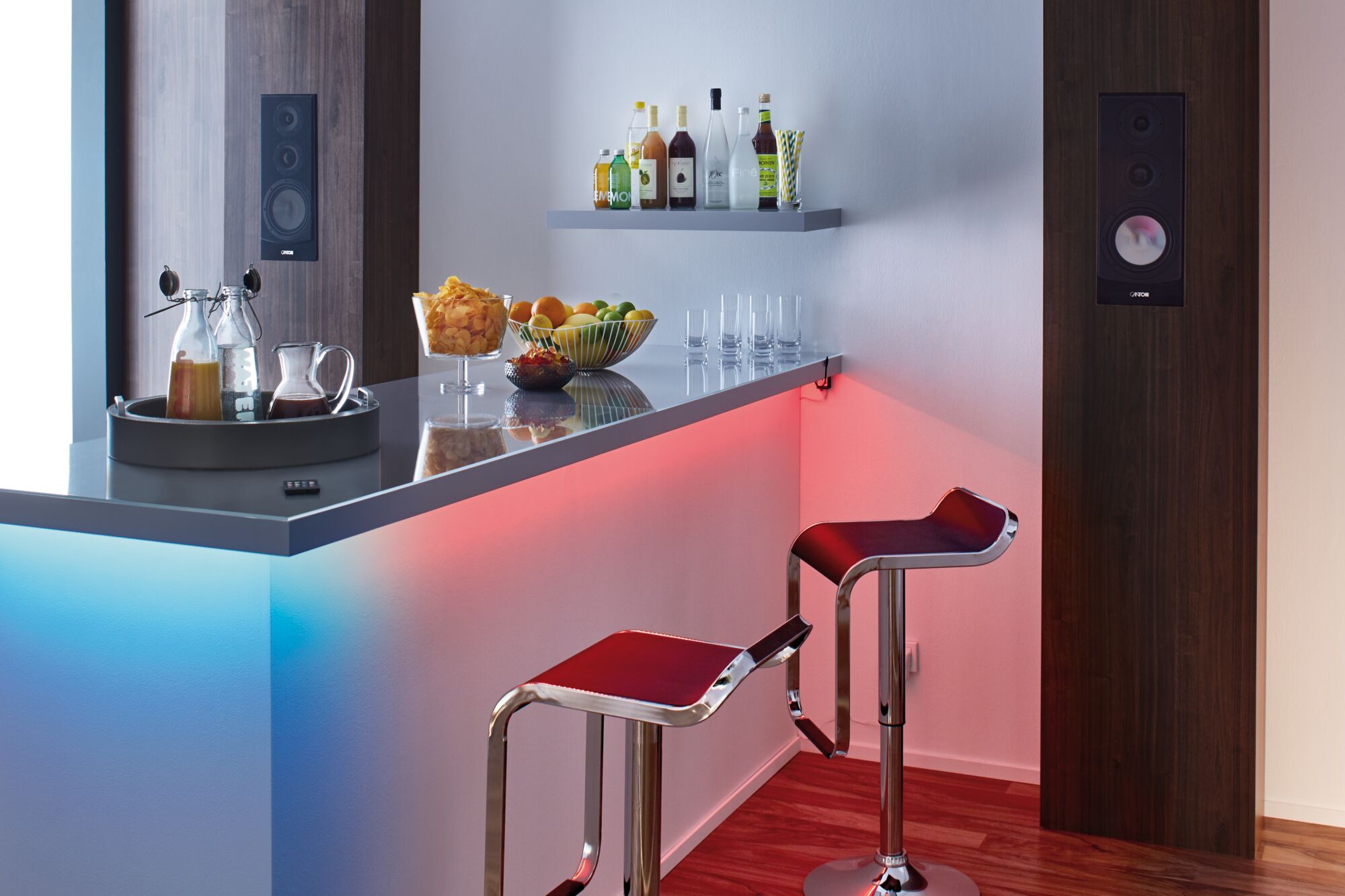 Moderne Bar mit LED-Beleuchtung in Blau und Rot, zwei roten Barhockern und Holzlautsprechern an der Wand