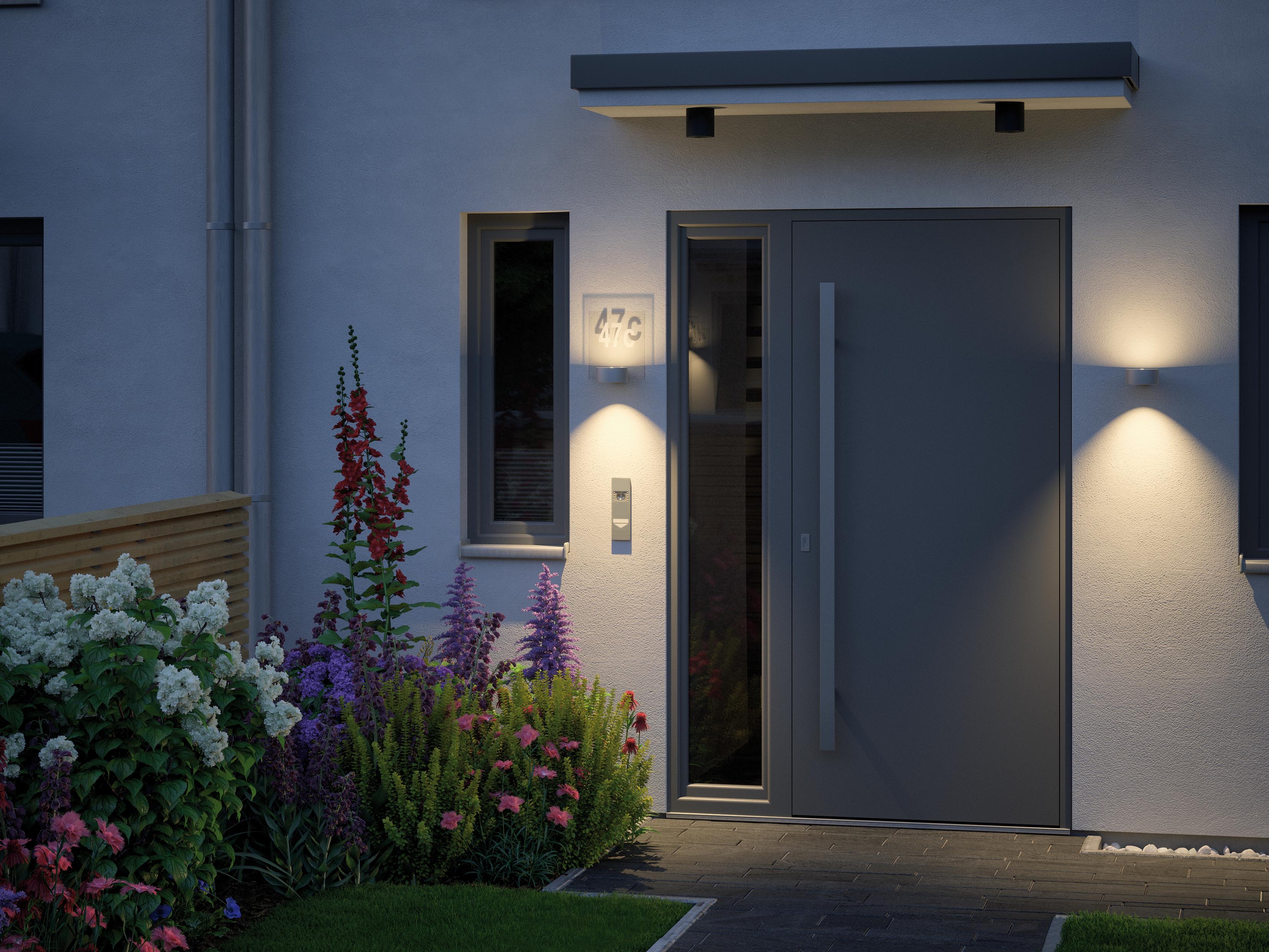 Moderner Hauseingang mit grauer Tür, beleuchtet von zwei Wandlampen und umgeben von bunten Blumen.