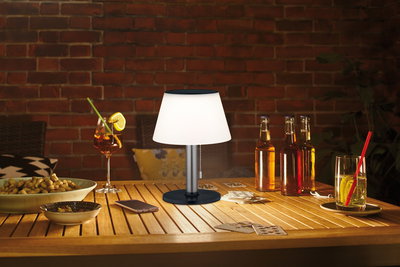 Moderne Tischlampe mit weißem Schirm und schwarzem Sockel auf Holztisch mit Getränken und Karten.