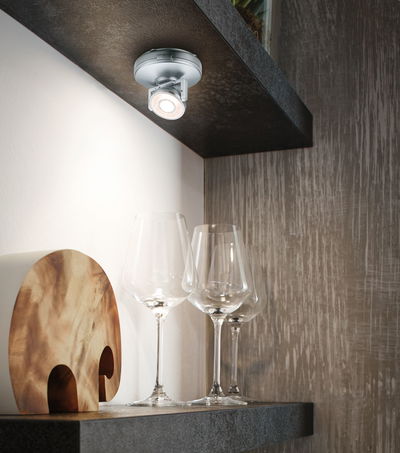 Spot LED de plafond argenté moderne éclairant trois verres à vin transparents sur étagère sombre.