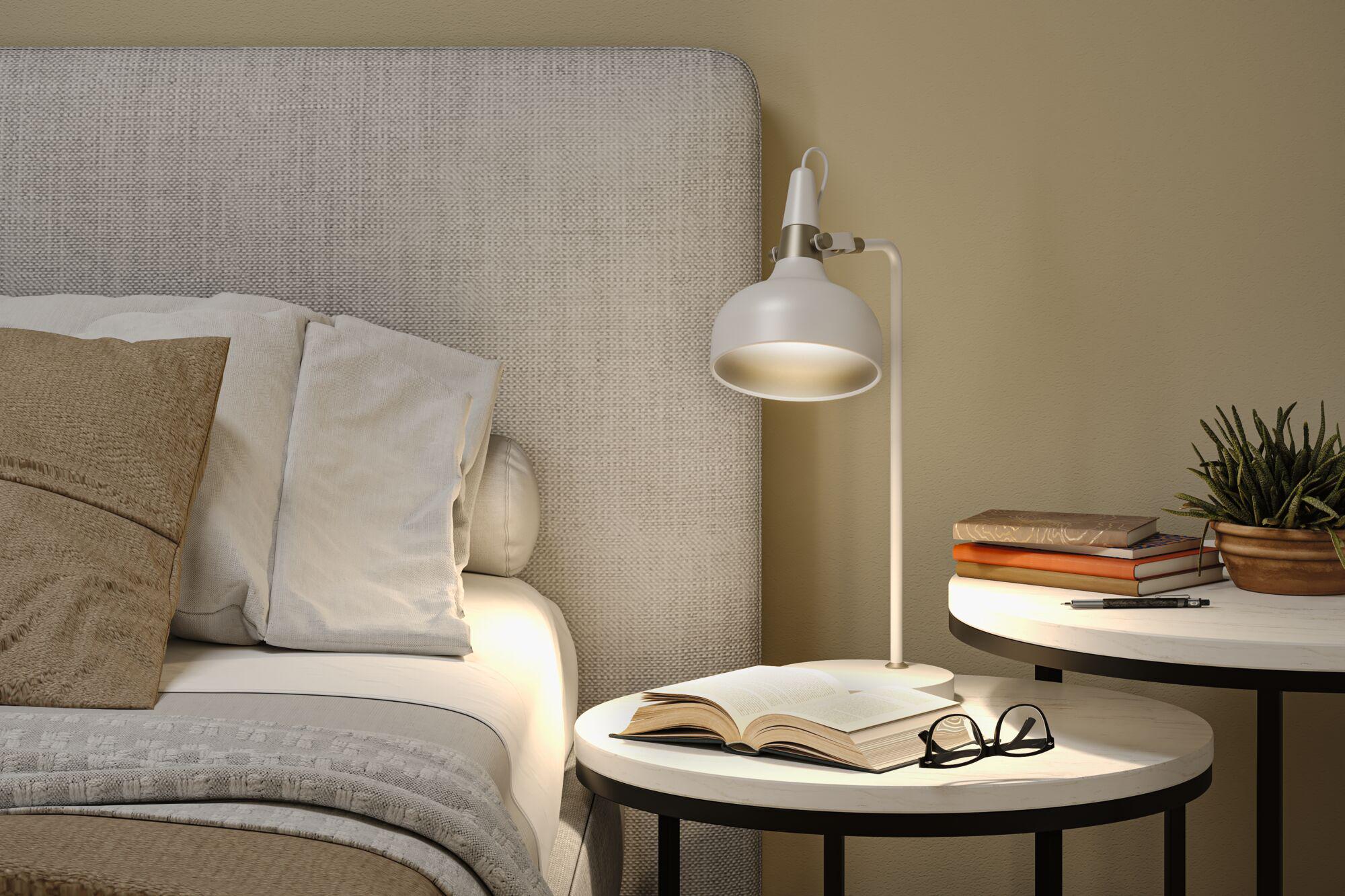 White metal floor lamp beside bed with beige-grey cushions and bedside table with books and plant